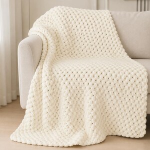 May include: Cream-colored, textured throw blanket draped over a light-colored sofa. The blanket features a raised, bobble-like pattern, creating a soft and inviting appearance. The neutral color and plush texture suggest comfort and warmth, suitable for home decor.