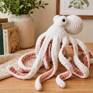 Könnte beinhalten: Ein handgefertigter weißer Häkel-Oktopus mit rosa Akzenten auf der Unterseite seiner Tentakel. Der Oktopus hat einen runden Kopf und acht lange, gebogene Tentakel. Der Oktopus befindet sich auf einer Holzoberfläche.