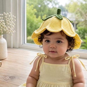 May include: A baby wearing a yellow crochet flower hat with a green stem and leaves. The baby is also wearing a yellow sundress with tie straps. The background includes a window and a vase of white flowers.