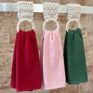 May include: Three kitchen towels in red, pink, and green hang from a metal bar. Each towel is held by a wooden ring and a crocheted holder in a neutral color. The towels have a textured appearance.