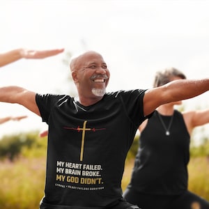 May include: A black t-shirt with a gold cross and the text "MY HEART FAILED. MY BRAIN BROKE. MY GOD DIDN'T." is worn by a smiling person in a yoga pose. Other people are in the background, also in yoga poses.