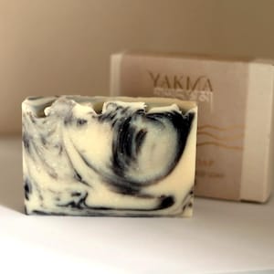 May include: A rectangular bar of soap with a marbled pattern of cream and black swirls. Behind the soap sits a beige cardboard box with the text YAKIMA printed in dark lettering, accompanied by a small graphic of wavy lines below the brand name.