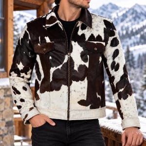 May include: A long-sleeve jacket featuring a brown and white cow print pattern. The jacket includes a pointed collar, a full-length front zipper closure, and two chest pockets with flaps secured by metallic buttons at the front of the garment.