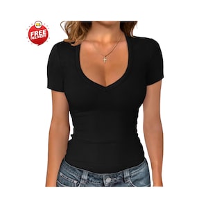 May include: Black short-sleeve V-neck shirt. The shirt has a ribbed texture and is worn with blue jeans. A gold-colored necklace with a cross pendant is visible. A red and white "FREE DELIVERY" sticker is in the upper left corner.