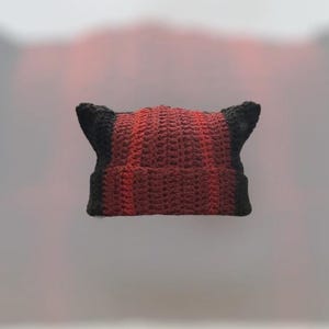 Op de afbeelding: Een handgemaakte gehaakte beanie in een uniek ontwerp. De hoed heeft een centraal gedeelte in roodtinten, dat overgaat in zwarte oorflappen. Een gezellig en stijlvol accessoire.