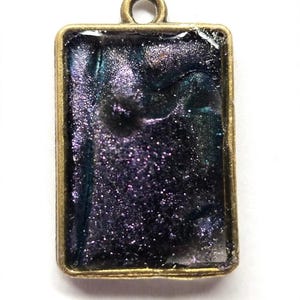 May include: A rectangular pendant featuring a metallic bronze-toned frame surrounding a dark, swirling resin inlay. The inlay displays a deep purple and black color palette with a fine, shimmering glitter finish throughout the surface.