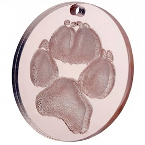 Puede incluir: Un colgante circular transparente con un diseño de huella de pata. El colgante es de color rosa claro y tiene un pequeño agujero en la parte superior para una cadena. La huella de la pata está grabada en la superficie.
