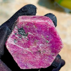 May include: A raw, hexagonal ruby crystal with a deep magenta and pink hue. The surface of the gemstone displays a natural, textured appearance with visible inclusions and dark mineral deposits embedded within the crystalline structure.