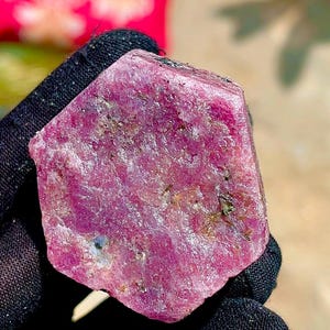 May include: A raw, hexagonal-shaped ruby crystal with a deep magenta and pink hue. The surface of the gemstone displays a natural, textured, and slightly translucent appearance with visible mineral inclusions throughout the crystalline structure.