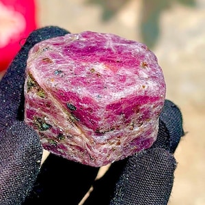 May include: A raw, hexagonal ruby crystal with a deep magenta and pink hue. The surface of the gemstone is textured and opaque, featuring natural inclusions and dark mineral deposits embedded within the crystalline structure of the stone.