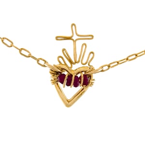 Puede incluir: Un collar de tono dorado con un colgante en forma de corazón con una cruz en la parte superior. El corazón está construido con alambre y adornado con tres pequeñas cuentas facetadas de color rojo oscuro envueltas horizontalmente en el centro.