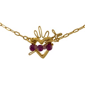 May include: A gold-toned necklace featuring a central heart-shaped pendant crafted from wire. The pendant includes a flame-like top, a small arrow detail, and three faceted deep pink beads strung horizontally across the center of the heart.