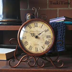 Könnte beinhalten: Eine runde Tischuhr mit einem kupferfarbenen Metallrahmen und einem dekorativen Sockel mit Schnörkeln. Das Zifferblatt hat einen cremefarbenen Hintergrund mit schwarzen römischen Ziffern, dem Text Winston Clock Co. Est. 1845 und dem Wort Quartz.