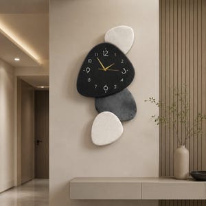 Peut inclure: Une horloge murale au design irrégulier en forme de galets, composée de quatre formes arrondies superposées dans des tons noir, gris foncé et blanc. La section centrale noire affiche des chiffres blancs de 1 à 12 et des aiguilles dorées.
