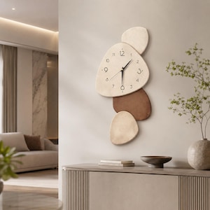 Peut inclure: Une horloge murale composée de trois formes irrégulières en galet qui se chevauchent, dans des tons crème et marron. La forme supérieure, la plus grande, présente des chiffres noirs de 1 à 12 et des aiguilles noires. Finition mate lisse.