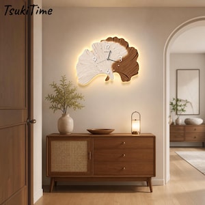 May include: A ginkgo leaf-shaped wall clock with a split design featuring a textured cream-colored side and a dark wood-toned side. The clock face includes white numbers at the 12, 3, 6, and 9 positions, black hands, and a warm integrated backlight.