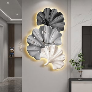 May include: A modern wall clock with a unique design. The clock features three stylized leaf shapes in shades of gray and white, with a warm yellow backlight. The clock hands are black, and the numbers are visible.