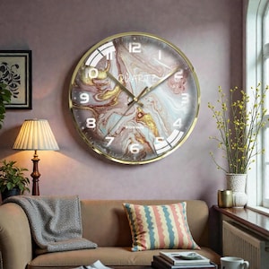 Pode incluir: Um relógio de parede redondo com uma moldura dourada. O mostrador do relógio apresenta um design de mármore em tons de rosa, dourado e branco. Os ponteiros e os números do relógio são dourados. A palavra "QUARTZ" está impressa no mostrador.