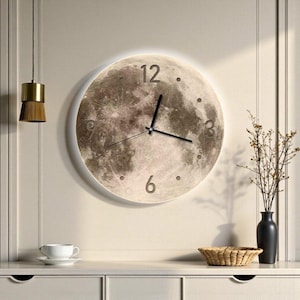 May include: A circular wall clock featuring a detailed lunar surface print with a soft glowing backlight. The clock face displays dark brown and beige craters, black hour and minute hands, and the numbers 3, 6, 9, and 12 in a simple font.