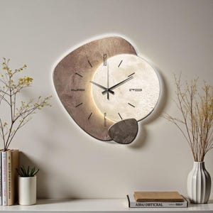 Puede incluir: Un reloj de pared moderno con un diseño único. El reloj presenta una esfera circular con una superficie texturizada de color blanquecino, rodeada por una forma abstracta marrón. Las manecillas del reloj son negras y el reloj está iluminado con una luz suave y cálida.