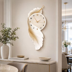 May include: A wall clock featuring a cream-colored circular face with black numerals and gold hands, integrated into a large, sculptural white feather design with gold accents that extends vertically along the wall behind the clock face.