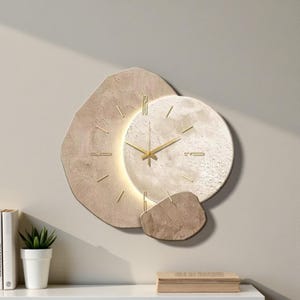 Puede incluir: Un reloj de pared moderno con un diseño único. El reloj presenta una esfera circular con manecillas y marcadores dorados, rodeada de formas texturizadas de color beige. El reloj está montado en una pared de color claro.