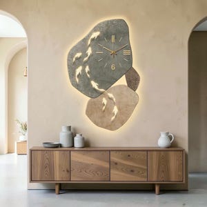 Puede incluir: Un reloj de pared con dos formas irregulares similares a piedras en gris y beige, iluminadas por una luz de fondo suave. La esfera muestra manecillas y marcadores dorados, con siluetas de peces koi dorados sobre las superficies.