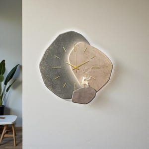 May include: A modern wall clock with a unique design. The clock features a layered, abstract shape in shades of gray and tan, with gold-colored hands and hour markers. A decorative deer silhouette is placed on the clock face.