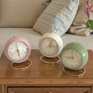 Puede incluir: Tres relojes analógicos esféricos en colores pastel: rosa, blanco y verde. Cada reloj tiene una base y manecillas doradas, con una esfera blanca y números negros. Los relojes se exhiben sobre una superficie de madera.