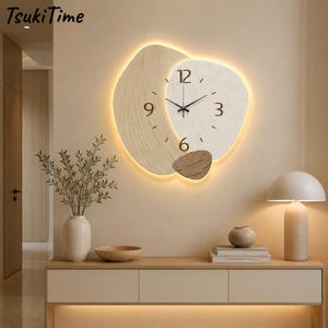 Könnte beinhalten: Eine Wanduhr mit überlappenden organischen Formen in heller Holzmaserung und strukturierter cremeweißer Oberfläche. Das Zifferblatt zeigt schwarze Zeiger und die Ziffern 3, 6, 9 und 12, wobei eine warme LED-Hintergrundbeleuchtung die Ränder betont.