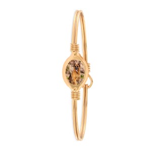 May include: Gold-colored bangle bracelet featuring an oval pendant with a photo of a German Shepherd dog. The bracelet has a wire-wrapped design and a hinged closure. The dog is brown and black.