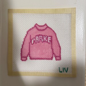 Op de afbeelding: Een kruissteekontwerp met een roze trui met de naam "PARKE" in witte letters. Het ontwerp staat op een wit gaasdoek met een beige rand. Het woord "LIV" is groen geborduurd aan de onderkant.