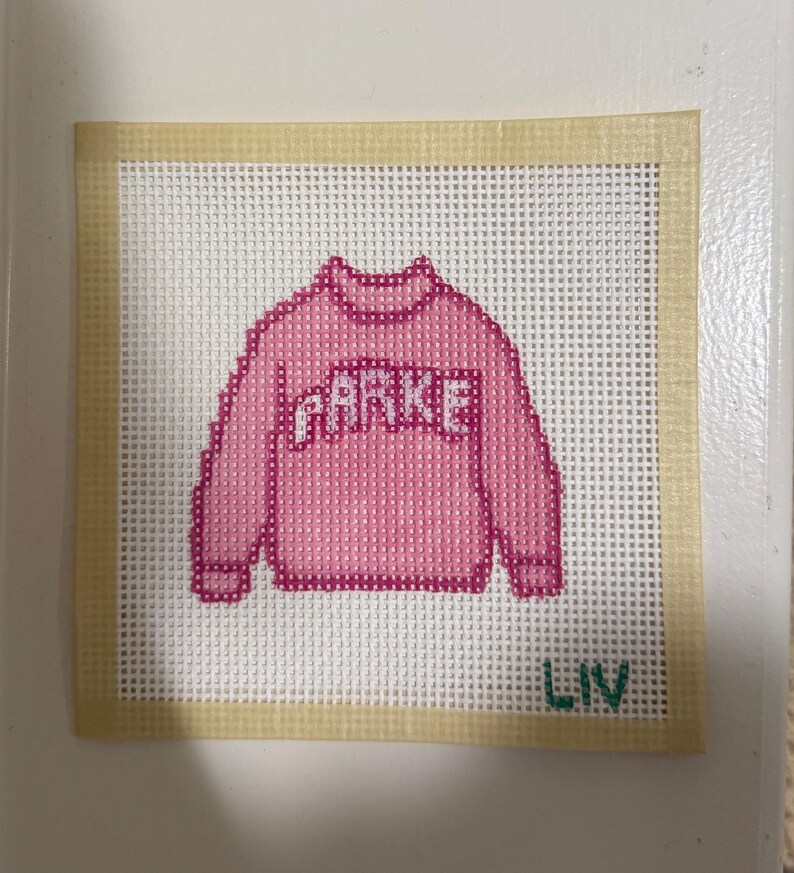 Op de afbeelding: Een kruissteekontwerp met een roze trui met de naam "PARKE" in witte letters. Het ontwerp staat op een wit gaasdoek met een beige rand. Het woord "LIV" is groen geborduurd aan de onderkant.