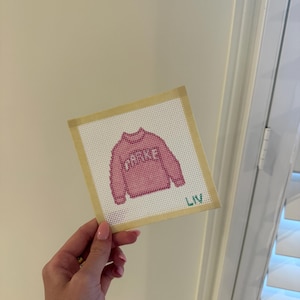 Op de afbeelding: Een roze kruissteekontwerp van een trui met het woord "PARKE" in witte letters. Het ontwerp staat op een wit canvas met een beige rand. De naam "LIV" is in groen rechtsonder geborduurd.