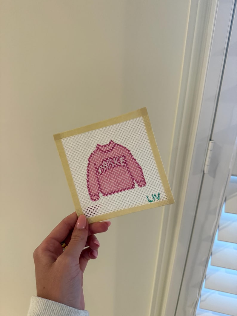 Op de afbeelding: Een roze kruissteekontwerp van een trui met het woord "PARKE" in witte letters. Het ontwerp staat op een wit canvas met een beige rand. De naam "LIV" is in groen rechtsonder geborduurd.