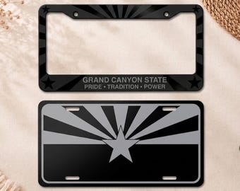 May include: A black license plate frame featuring a sunburst pattern, two stars, and the text GRAND CANYON STATE PRIDE TRADITION POWER, paired with a rectangular license plate displaying a grey and black sunburst design with a central star.