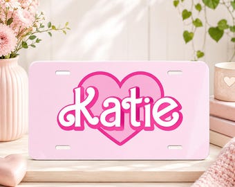 May include: A rectangular light pink license plate featuring a large, dark pink heart graphic in the center. The name Katie is printed in a bold, white, stylized script font with a dark pink outline across the middle of the heart shape.