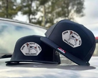 May include: Two navy blue trucker hats with a white patch featuring a hound dog and the words "COON HUNT WITH WALKERS". The hats have a mesh back and a flat brim. The hats are sitting on a car.