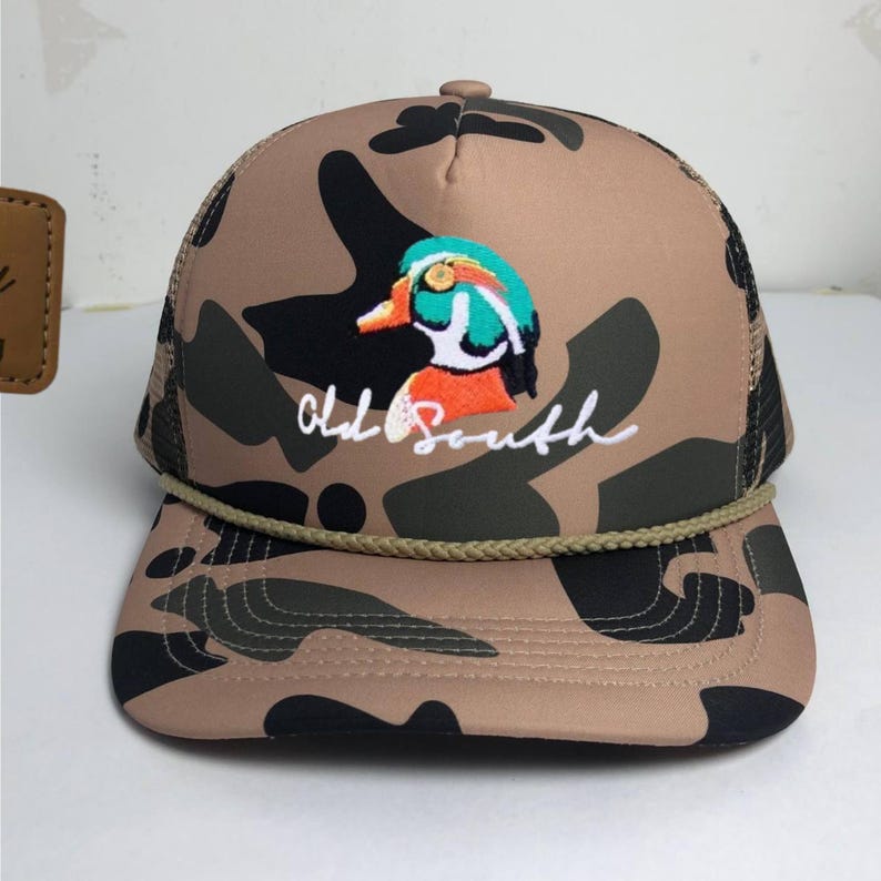 May include: A camouflage trucker hat with a brown, black, and green pattern. The hat features an embroidered duck design with teal, orange, and white accents, and the words "Old South" in white script.