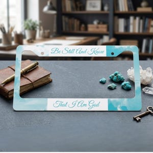 May include: A rectangular license plate frame featuring a light blue sky and white cloud pattern. The top horizontal bar displays the text "Be Still And Know" in a teal script font, while the bottom bar displays "That I Am God" in the same font.