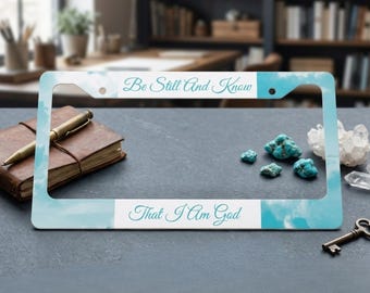 May include: A rectangular license plate frame featuring a light blue sky and white cloud pattern. The top horizontal bar displays the text "Be Still And Know" in a teal script font, while the bottom bar displays "That I Am God" in the same font.