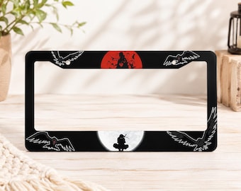 May include: A black rectangular license plate frame featuring anime-inspired graphics. The top center displays a red circular sun with a silhouette, while the bottom center shows a white full moon with a dark ninja figure and flying crows.