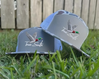 May include: Two gray trucker hats with a camouflage pattern on the bill and a light blue mesh back. Each hat features an embroidered flying duck and the text "True South" in white script.
