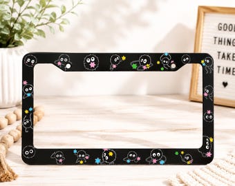 May include: A black rectangular license plate frame featuring a repeating pattern of small, round, black soot sprite characters with large white eyes. The sprites are depicted interacting with colorful, multi-pointed star shapes in pink, blue, and yellow.