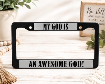 May include: A black license plate frame featuring two silver-toned horizontal inserts. The top insert displays the black text MY GOD IS, while the bottom insert features the black text AN AWESOME GOD! The frame includes four circular mounting holes.