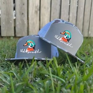 May include: Two gray trucker hats with a duck logo and the text "Old South." The hats have a camouflage pattern on the bill and a light blue mesh back. The duck logo features orange, teal, and white colors.