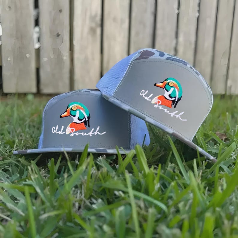 May include: Two gray trucker hats with a duck logo and the text "Old South." The hats have a camouflage pattern on the bill and a light blue mesh back. The duck logo features orange, teal, and white colors.