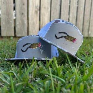 May include: Two gray trucker hats with a fish-like graphic and the initials "AF" embroidered on the side. The hats have a camouflage brim and a light blue mesh back. The hats are resting on green grass.