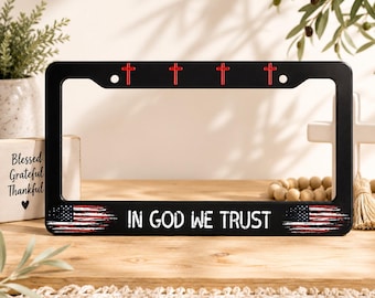 May include: A black license plate frame featuring four small red crosses along the top edge. The bottom section displays the text IN GOD WE TRUST in white capital letters, flanked by two distressed American flag graphics on the left and right sides.