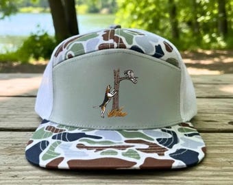 May include: A trucker hat with a camouflage pattern on the brim and top. The front panel is light gray with an illustration of a dog climbing a tree. The hat has a white mesh back.